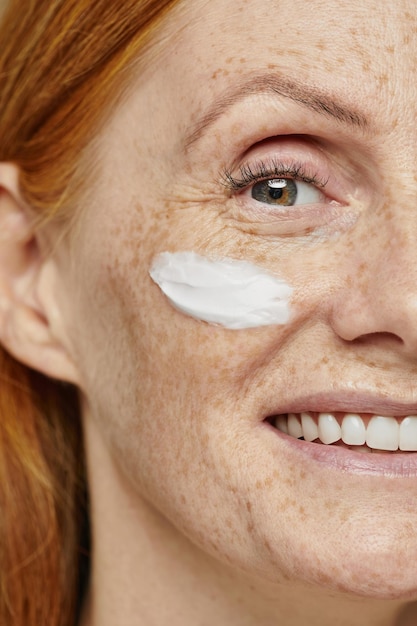 Close up half face portrait of freckled red haired woman using face cream and smiling at camera