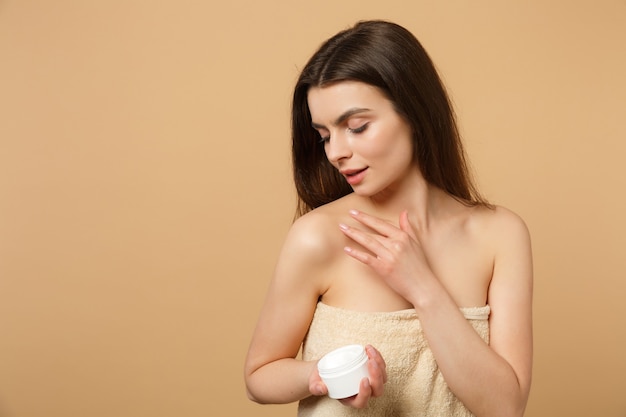 Close up half naked woman with perfect skin nude make up applying facial cream isolated on beige pastel wall