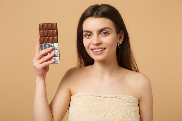 Close up half naked woman with perfect skin, nude make up holds chocolate bar isolated on beige pastel wall