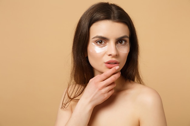 Close up half naked woman with perfect skin, nude make up patches under eyes isolated on beige pastel wall