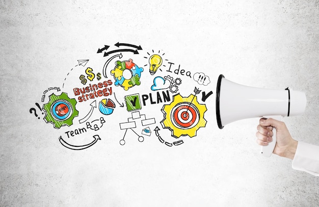 Close up of a hand of a buisnessman in a white shirt holding a megaphone near a concrete wall with a colorful business plan drawing