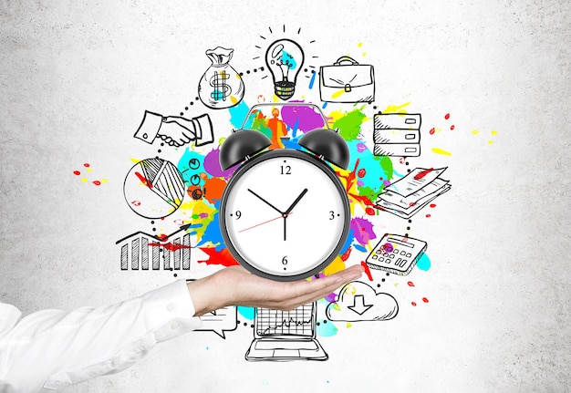 Close up of a hand of a businessman in a white shirt holding an alarm clock. Concrete background with time management icons