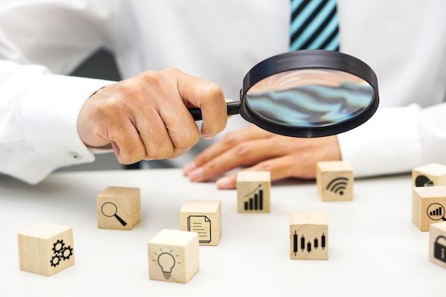 Close up hand of businessperson looking at wooden blocks with icon symbols through magnifying glass