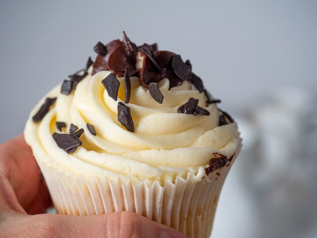 Close-up in the hand of a cupcake with white cream decorated with pieces of dark chocolate. Sweets, delicious dessert. Calorie content, unhealthy food