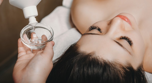 Close up hand of a female cosmetologist using a transparent mask from a bottle to apply on a female face for skin care treatment.