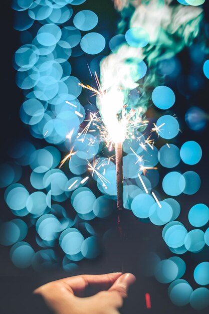 Photo close-up of hand holding burning sparkler against illuminated lights at night