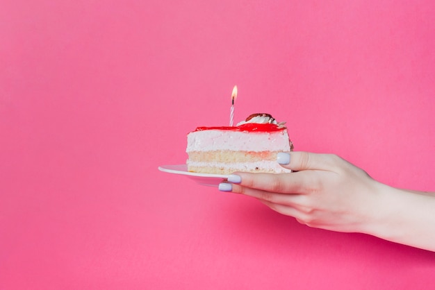 Close-up della mano che tiene la fetta di torta con candela accesa sul piatto sopra lo sfondo rosa