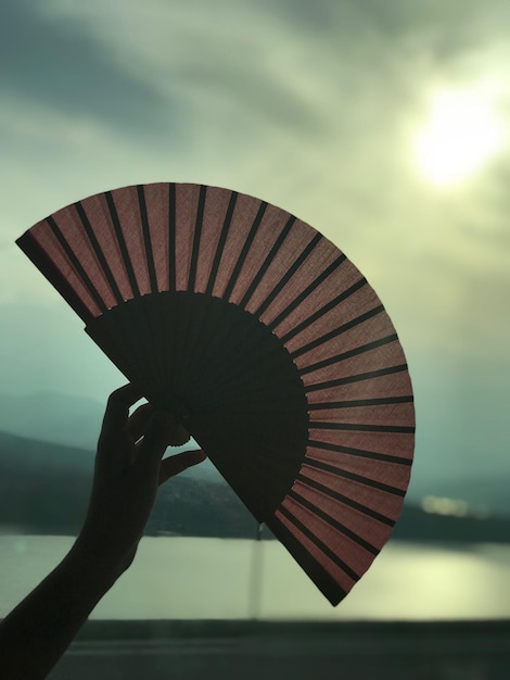 Photo close-up of hand holding folding fan against cloudy sky