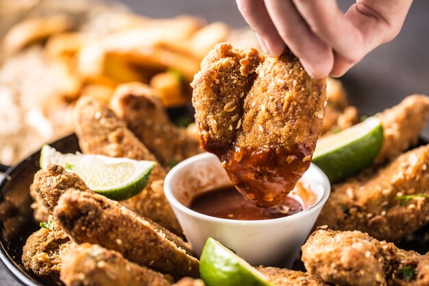 Close-up hand holding fried chicken wing with chili sauce.