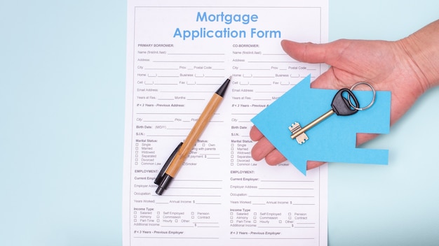 Close-up of a hand holding a key in a blue paper house over a home mortgage application and a pen, top view