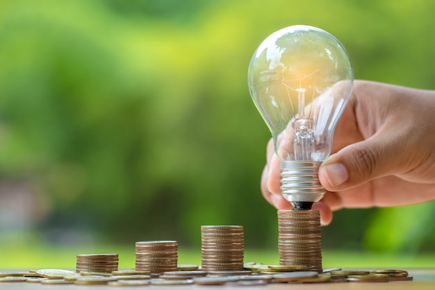 Close up hand holding light bulb with Coins stack on wooden table.