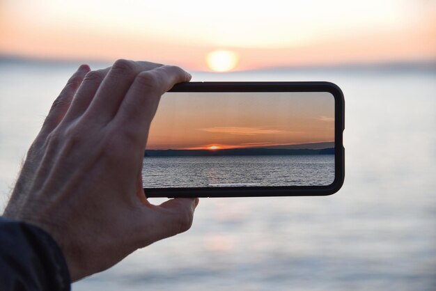 Photo close-up of hand holding mobile phone against sea during sunset