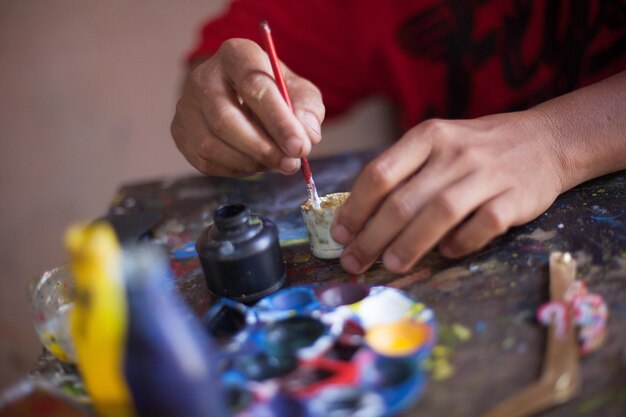 Photo close-up of hand holding painting brush at messy table