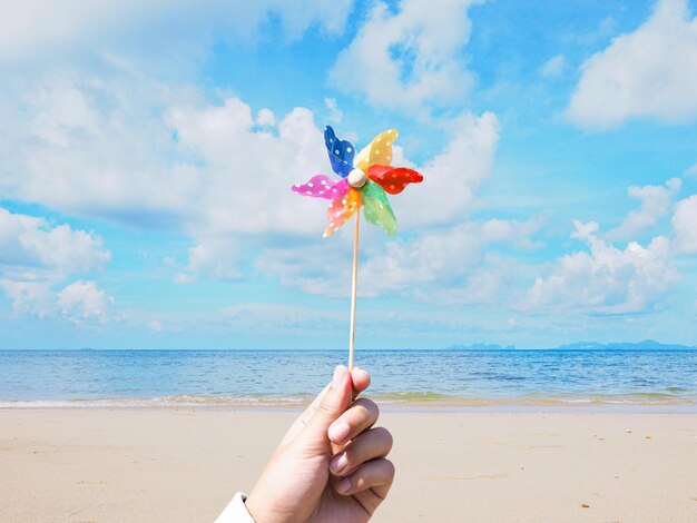 Foto primo piano della mano che tiene la girandola sullo sfondo della spiaggia estiva giocattolo colorato per bambini con turbina eolica