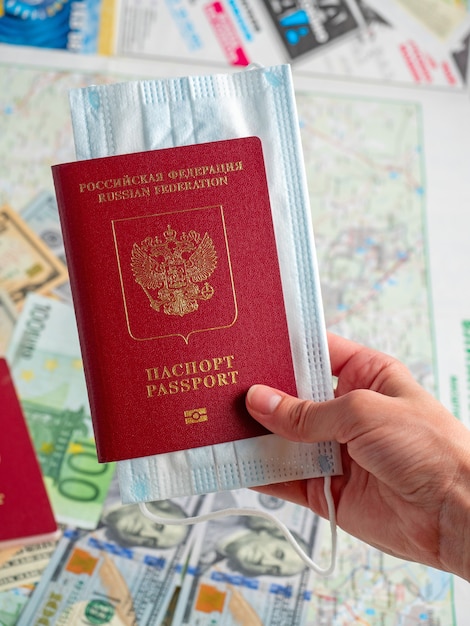 Close-up of a hand holding a red passport of the Russian Federation with a medical mask attached. The map is in the background. The concept of travel during a pandemic