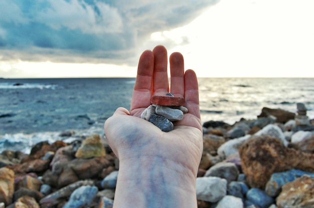 Foto close-up di una mano che tiene una roccia sulla spiaggia contro il cielo