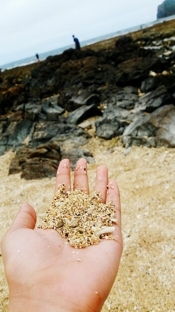 Photo close-up of hand holding rock on sand at beach against sky