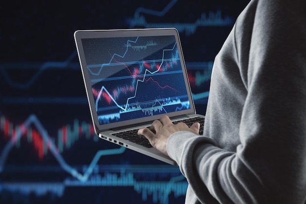 Close up of hand holding and using laptop with glowing forex index chartgraph on dark background Trade finance and fintech concept