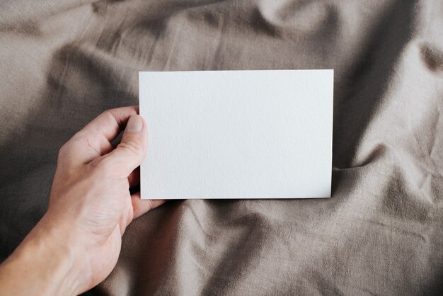 Close-up of hand holding white greeting card mock-up with empty space on rustic textile background