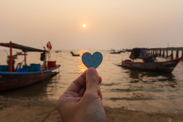 Foto close up di mano uomo che tiene un cuore di carta con lo sfondo del mare in serata concetto d'amore felice