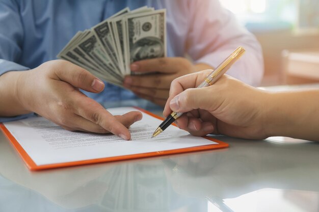 Photo close-up of hand signing documents with person holding bribe on table