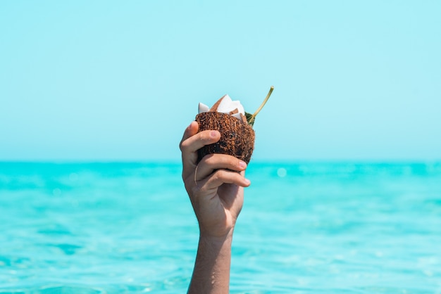 Close up of hand of an unrecognizable person holding half coconut shell with slices on coconut in it against sea and sky. Wet hand in summer with coconut shell. Raised hand holding coconut against sea