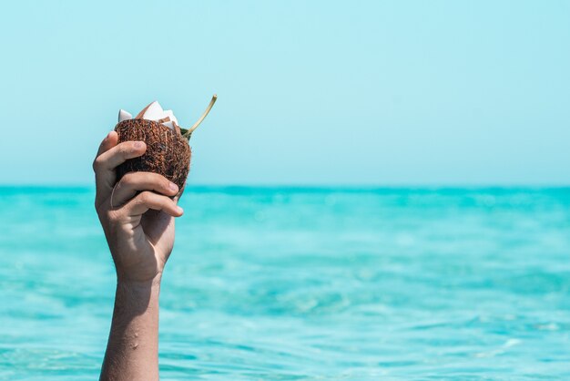Close up of hand of an unrecognizable person holding half coconut shell with slices on coconut in it against sea and sky. Wet hand in summer with coconut shell. Raised hand holding coconut against sea