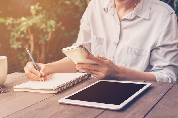 ノートを書くとヴィンテージトーンとコーヒーショップで電話を保持する手の女性を閉じます。