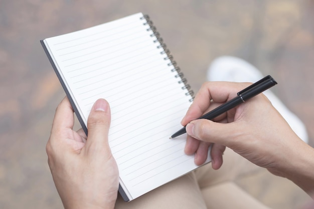 Close up hand young man are using pen writing Record Lecture note pad into the book sitting on the chair outdoor.