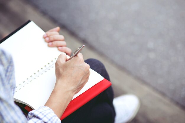 Close up hand young man are using pen writing Record Lecture note pad into the book sitting on the chair outdoor.