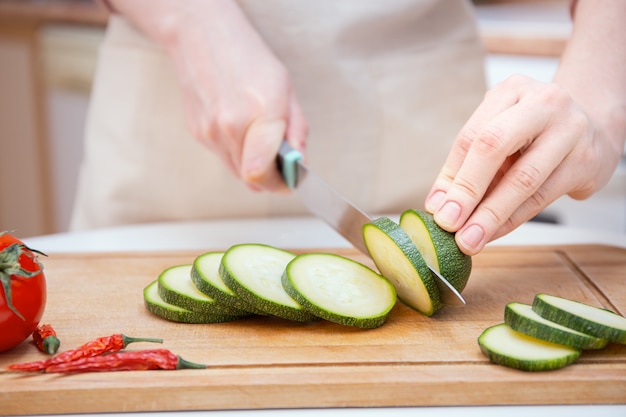 Close-up Handen van een jonge vrouw gesneden met een mes in plakjes of plakjes jonge courgette komkommer op een houten snijplank. Bereiding van ingrediënten en groenten voor het koken en grillen.