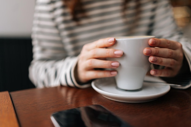 Close-up handen van onherkenbare jonge vrouw met een kop koffie die aan tafel zit met een mobiele telefoon tijdens een heerlijk ontbijt in een modern café Vrouwelijke handen met een kop koffie en een smartphone