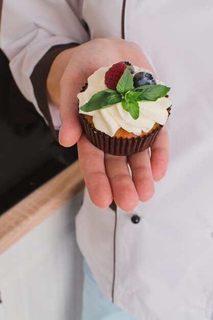 Foto primo piano sulle mani del pasticcere femminile in abito bianco con muffin o un cupcake decorato con frutti di bosco