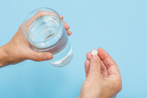 Close up hands of female patient with glass of water in one hand and white pill in another. Top view with blue background and copy space