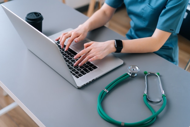 Close-up of hands of female practitioner doctor using typing on laptop keyboard sitting at desk 