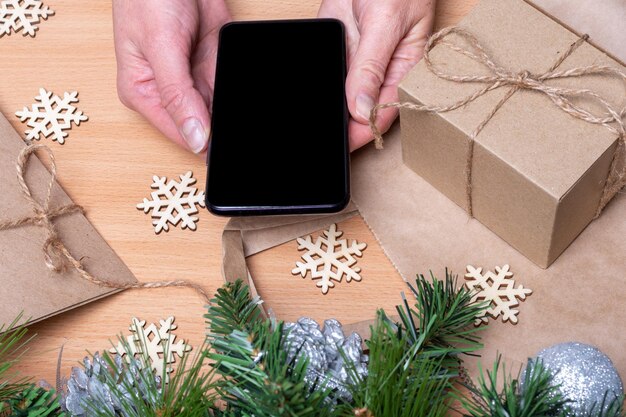 Close up hands holding mobile phone with blank black screen on wooden table with Christmas gift boxes, snowflakes, Christmas tree branches. Online shopping christmas gifts concept