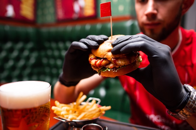 レタスとマヨネーズとおいしいグリルビーフバーガーを持っているクローズアップの手。ファーストフードで食べる若者のグループ。カフェ、ビールパブ、バーで一緒に時間を過ごす友達。