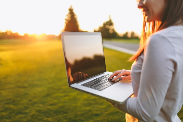 Close up hands on keyboard. Woman working on laptop pc computer with blank black empty screen to copy space in park on green grass sunshine lawn outdoors. Mobile Office. Freelance business concept.