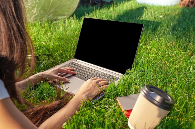 Close up hands on keyboard. Woman working on laptop pc computer with blank black empty screen to copy space in park on green grass sunshine lawn outdoors.