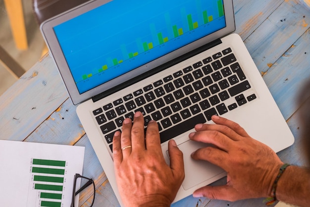 Photo close up of hands of man typing on his laptop and watching some stadistics of his work - blue wooden table