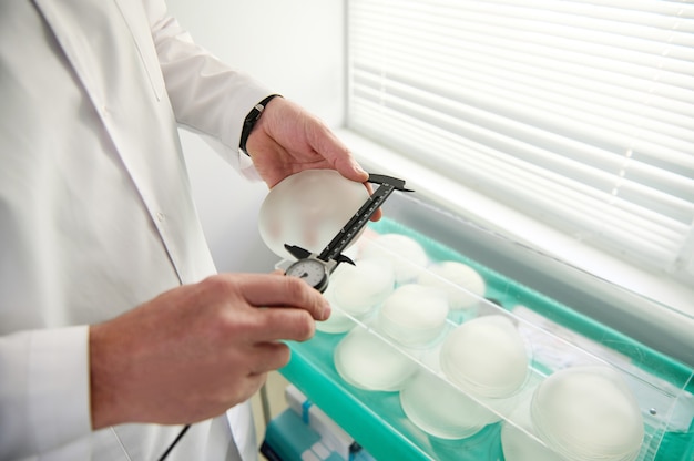 Close-up of hands of plastic surgeon holding a silicone breast implant and measuring its size with a calliper on the background of silicone implants of different shapes and sizes. Mammoplasty concept