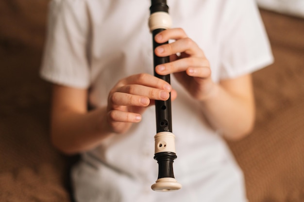 Close-up hands of talented adorable little girl playing flute sitting on bed in bedroom. Child practices playing musical instrument at home. Concept of leisure activity at home.