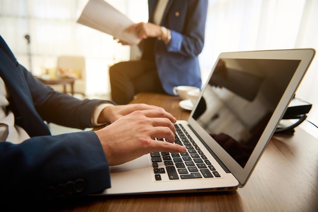 Close-up of the hands of unrecognizable man in business suit working on laptop, typing text on keyboard on the background of his female partner, colleague sitting at table with stack of documents