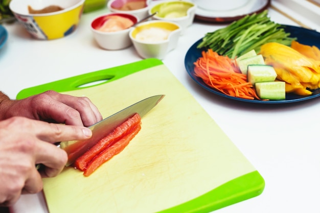 Photo close up of the hands of an unrecognizable person cutting raw and fresh salmon with a knife in a cutting board next to ingredients and sauces