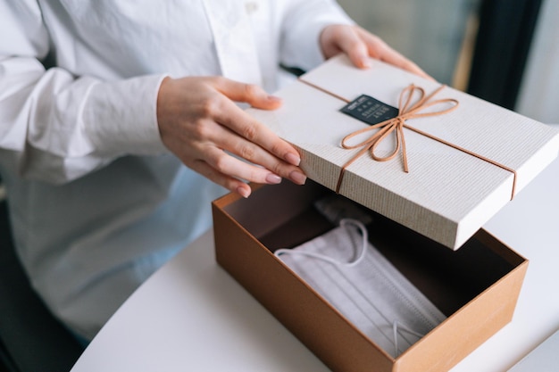 Mani ravvicinate di una donna irriconoscibile che aprono la confezione regalo ed estraggono la maschera protettiva medica alla scrivania.