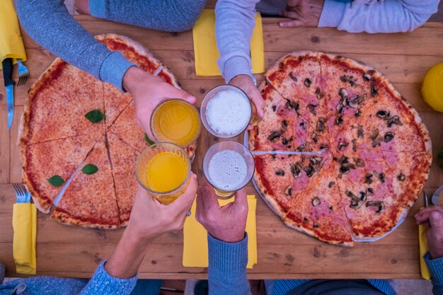 テーブルの中央でビールとオレンジジュースが一緒にチリンと鳴るガラスで手のクローズアップ-木製のテーブルの上の2つの大きなピザ