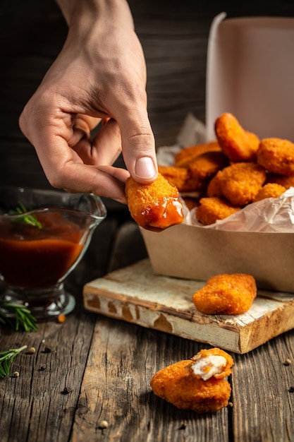 Крупный план рук с домашними вкусными куриными наггетсами и соусом в бумажной коробке на деревянной поверхности