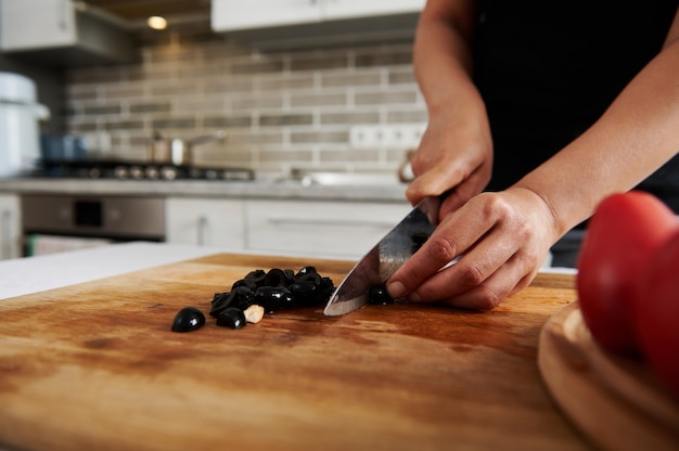 Close-up of hands of a woman chopping black olives on a wooden board. Food preparations concepts