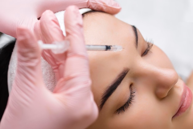 Close up of hands of young cosmetologist injecting botox in female face She is standing and smiling The woman is closed her eyes with relaxation