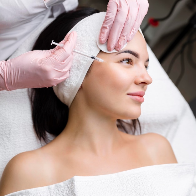 Photo close up of hands of young cosmetologist injecting botox in female face she is standing and smiling the woman is closed her eyes with relaxation